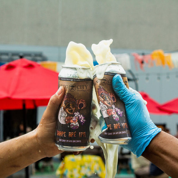 Two cans of Grape Ape IPA being smashed together and foamy beer pouring out. One hand wearing a blue latex glove.