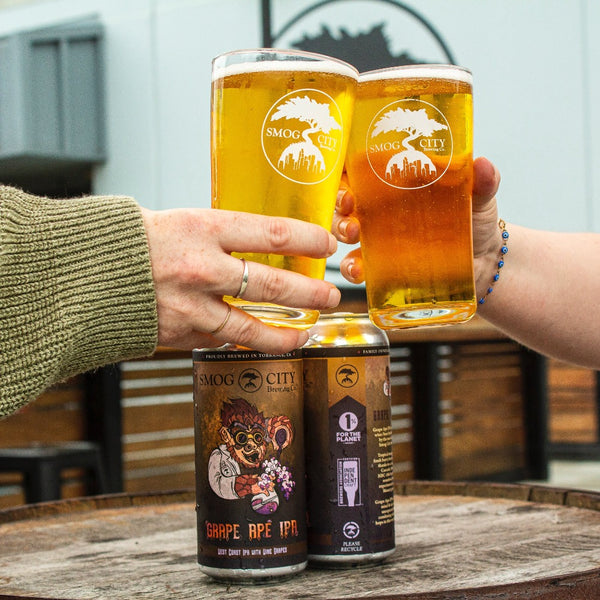 two hand cheersing glasses of beer above two cans of Grape Ape IPA on a wooden abrrel