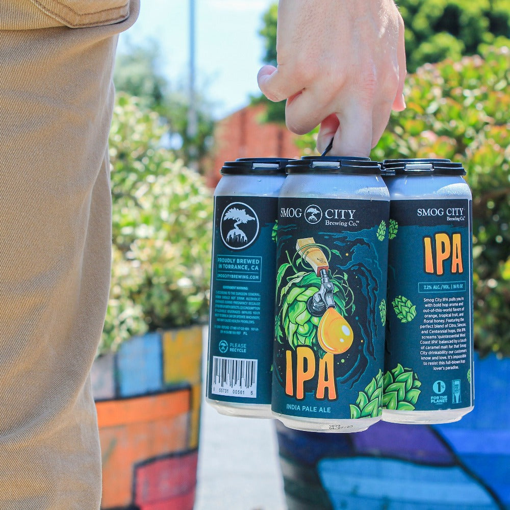 person holding a 4-pack of Smog City IPA at knee height wearing tan jeans, colorfully painted planters and bushes in the background.