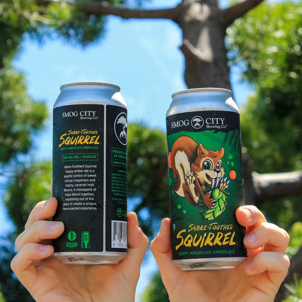 two hands holding cans of sabre-toothed squirrel, trees and blue sky in the background