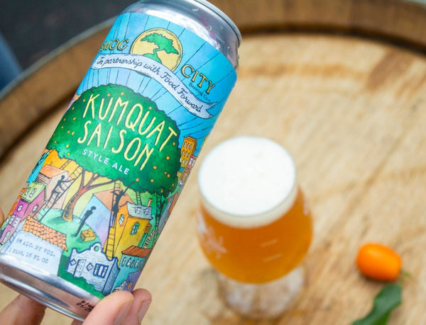 can of kumquat saison held above a wooden barrel with tulip glass of the beer and a kumquat fruit in the background.