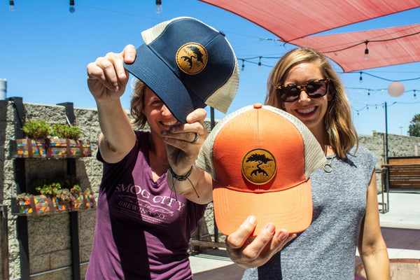 Two women holding blue and orange smog city trucker hats towards the camera
