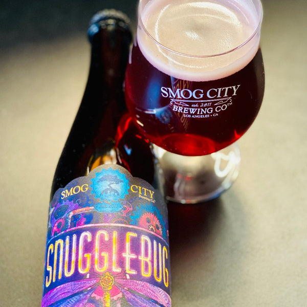 bottle of snugglebug lying down next to a tulip beer glass of snugglebug sour. Bright red beer with a creamy off white head.